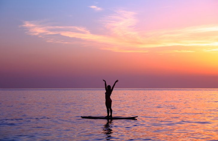 夕焼けの波のないビーチでサーフボードに乗って伸びをしている女性のシルエット