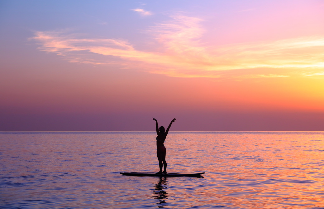 夕焼けの波のないビーチでサーフボードに乗って伸びをしている女性のシルエット