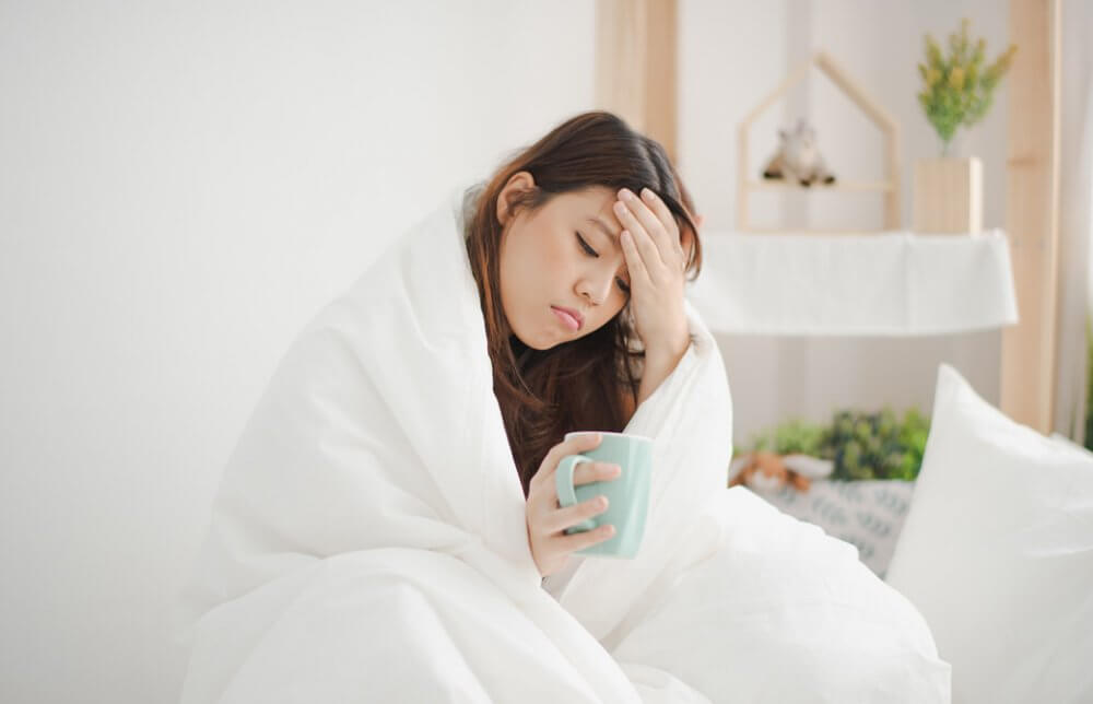 ベッドの上で毛布にくるまりおでこをおさえて具合悪そうにコーヒーカップを手にする女性