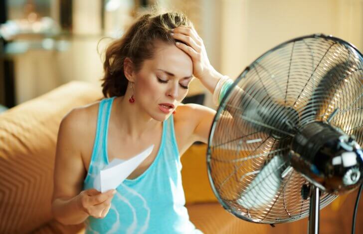扇風機の前で暑そうに顔を扇いで頭を抱えている女性