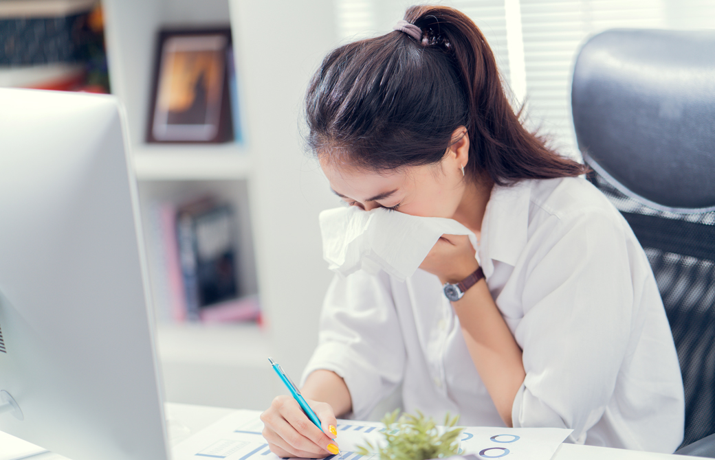 デスクで仕事中の女性がくしゃみを手でおさえている Azumi Shutterstock ヨガジェネレーション Yogageneration