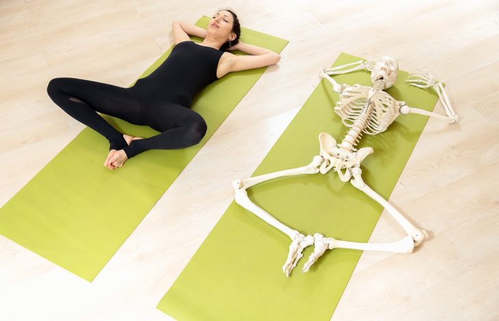 ヨガマットの上でスプタバッタコナーサナをする女性と同じ姿勢の骨模型