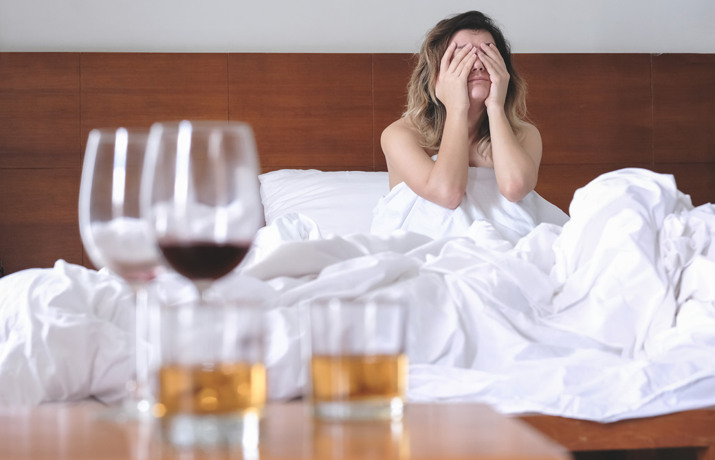 二日酔いでベッドの上で顔を覆っている女性とテーブルの上の飲み残しのお酒のグラス
