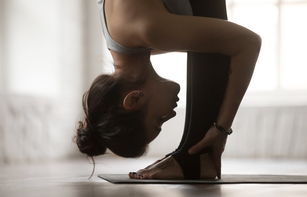室内でヨガマットの上で立位前屈をしている女性を横から見たところ
