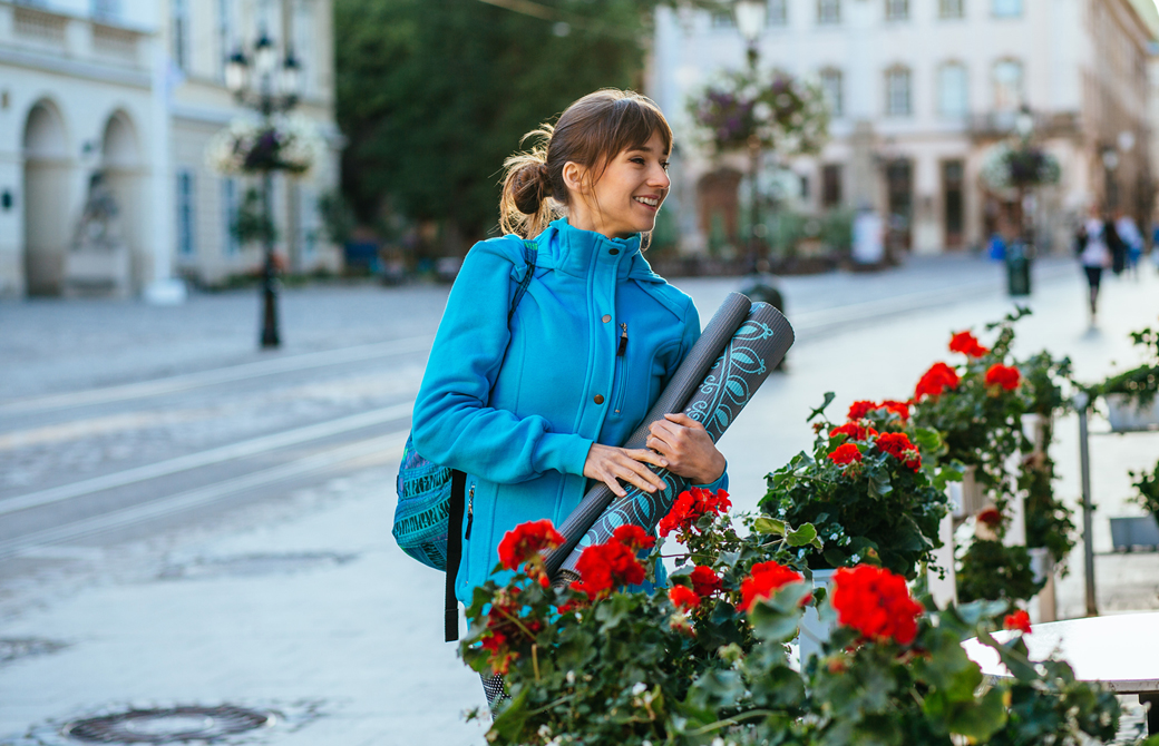 ヨガマットを持った青いジャケットを着ている女性がバラの生け垣の前で微笑んでいる