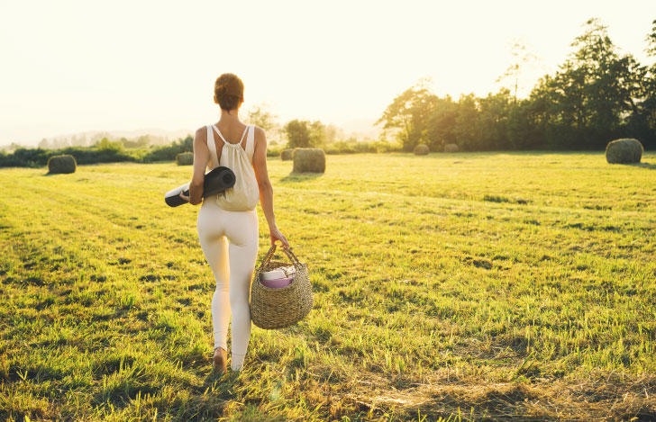 芝生の中を歩くヨガマットを抱えリュックサックを背負い手には大きなバッグを持った女性の後ろ姿