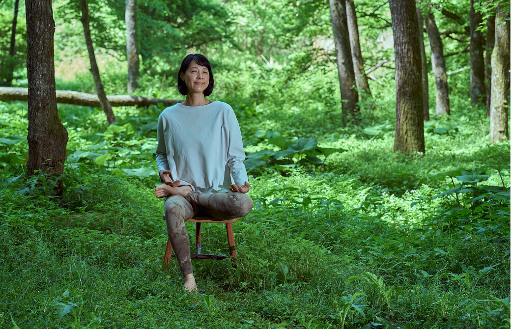 森の中で椅子に座って片足を反対の腿の上に載せて微笑んでいる山田いずみ先生
