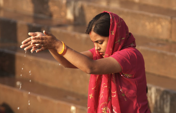 ガンジス川で両手で水を掬い上げる赤い布を纏った女性