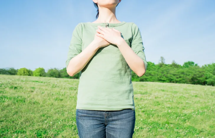 芝生と青々としげる木々を背景に胸に手を当てるカジュアルな服装の女性