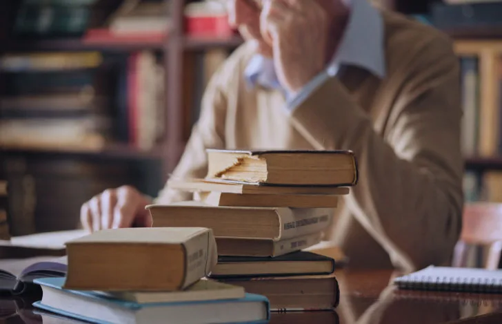 書斎の中の本が積み上がった机に座る男性の姿