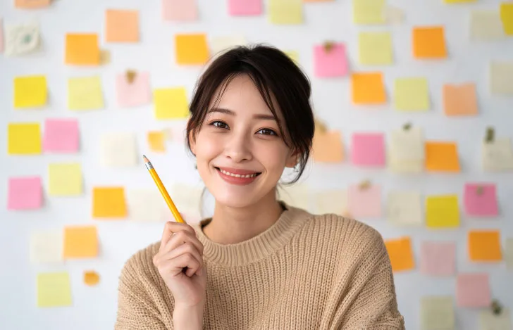 付箋が貼られた壁の前で鉛筆を持って微笑む女性